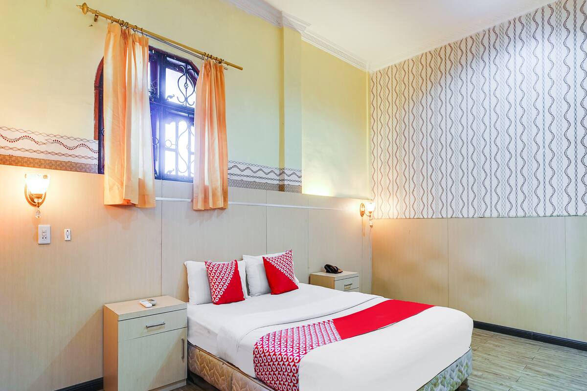 Ein Hotelzimmer mit Bett, Nachttischen, einem Fenster mit Vorhängen, Wandleuchten und ornamentalem Wandpapier.