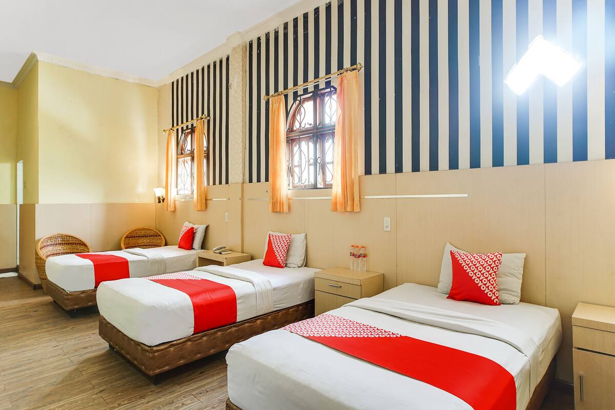 Ein Hotelzimmer mit drei Betten, jeweils mit weißer Bettwäsche und roten Akzenten, Holz-Kopfteilen und einer gestreiften Wand.