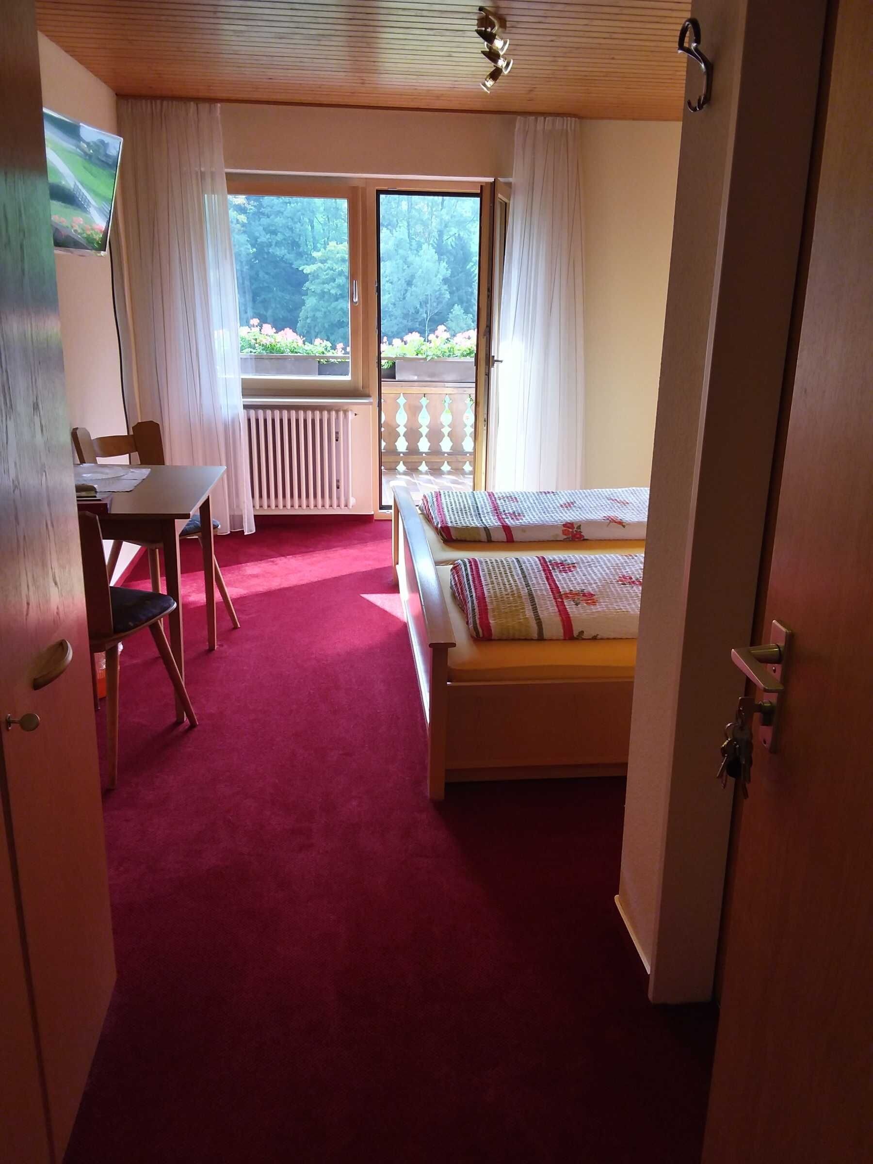 Ein Schlafzimmer mit einem Bett, einem Schreibtisch und einem Stuhl. Ein Fenster mit Vorhängen bietet Blick auf Grünfläche.