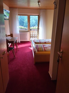 A bedroom with a bed, a desk, and a chair. There is a window with curtains and a view of greenery outside. - Double room 2 with toilet and shower, 1 - 2 persons - Mosertonihof (Elzach)