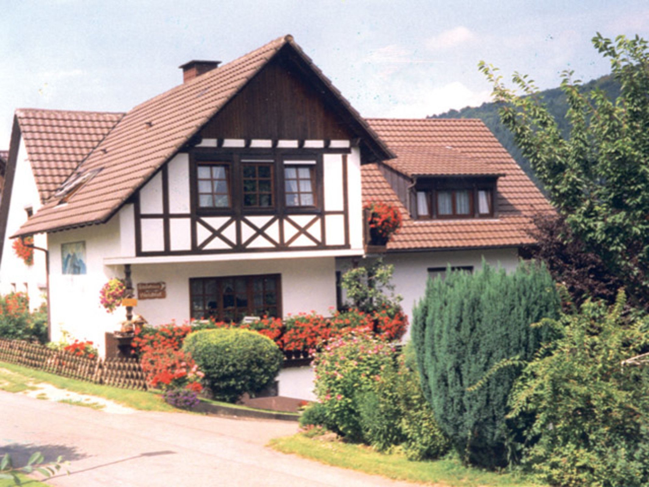 Une maison traditionnelle à colombages, dotée d’un toit à deux versants, d’un porche à l’avant et d’un jardin bien entretenu où poussent diverses plantes et fleurs.