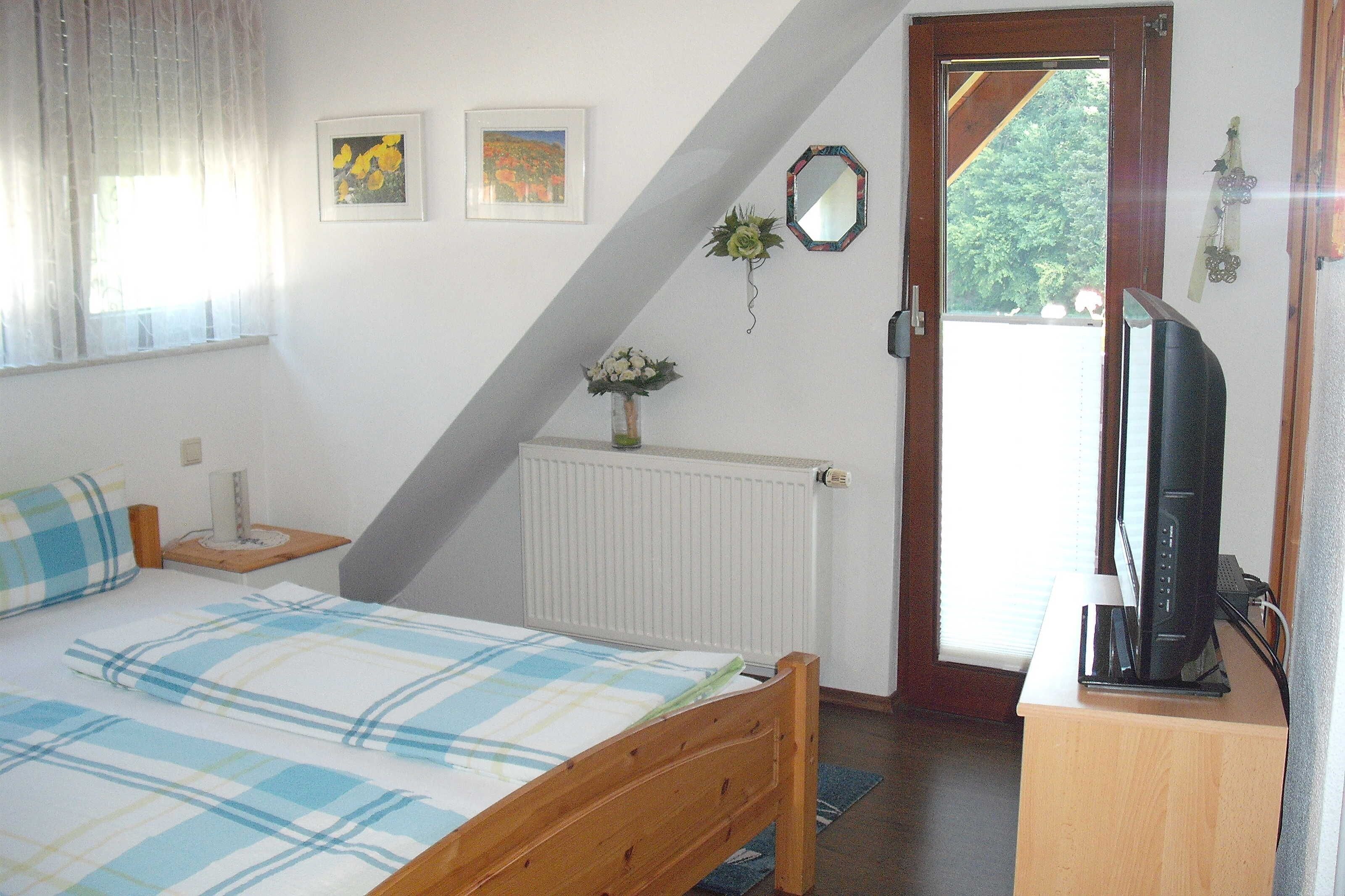 Ein Schlafzimmer mit einem Bett, einem Schreibtisch mit Computer, einem Fenster mit Vorhängen und einer Tür, die nach draußen führt.