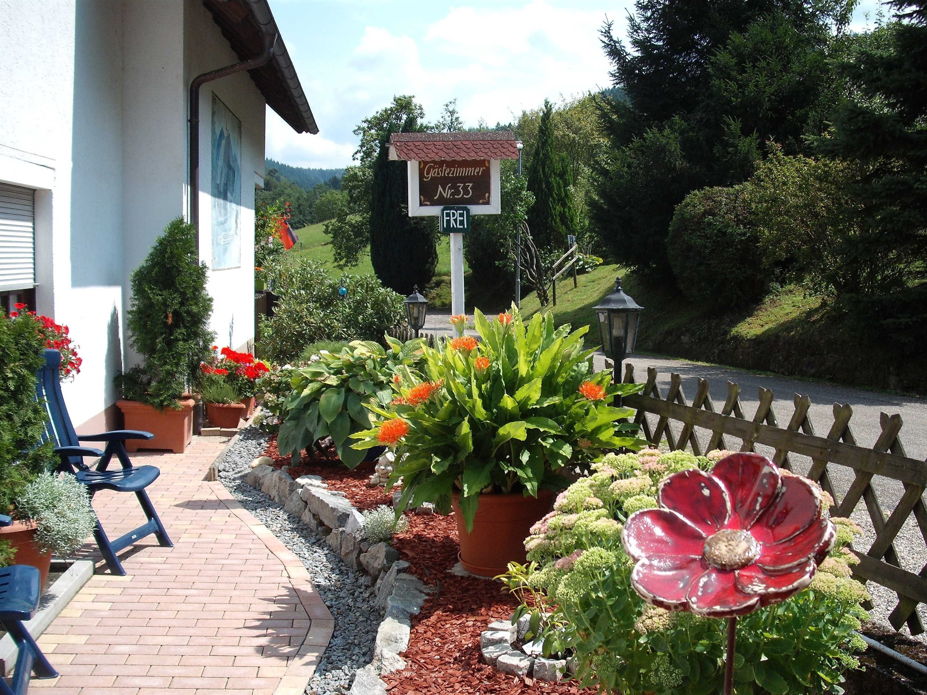Un bâtiment avec une enseigne indiquant qu’il s’agit d’une maison d’hôtes, un chemin piétonnier dallé et une variété de plantes et de fleurs en pot.