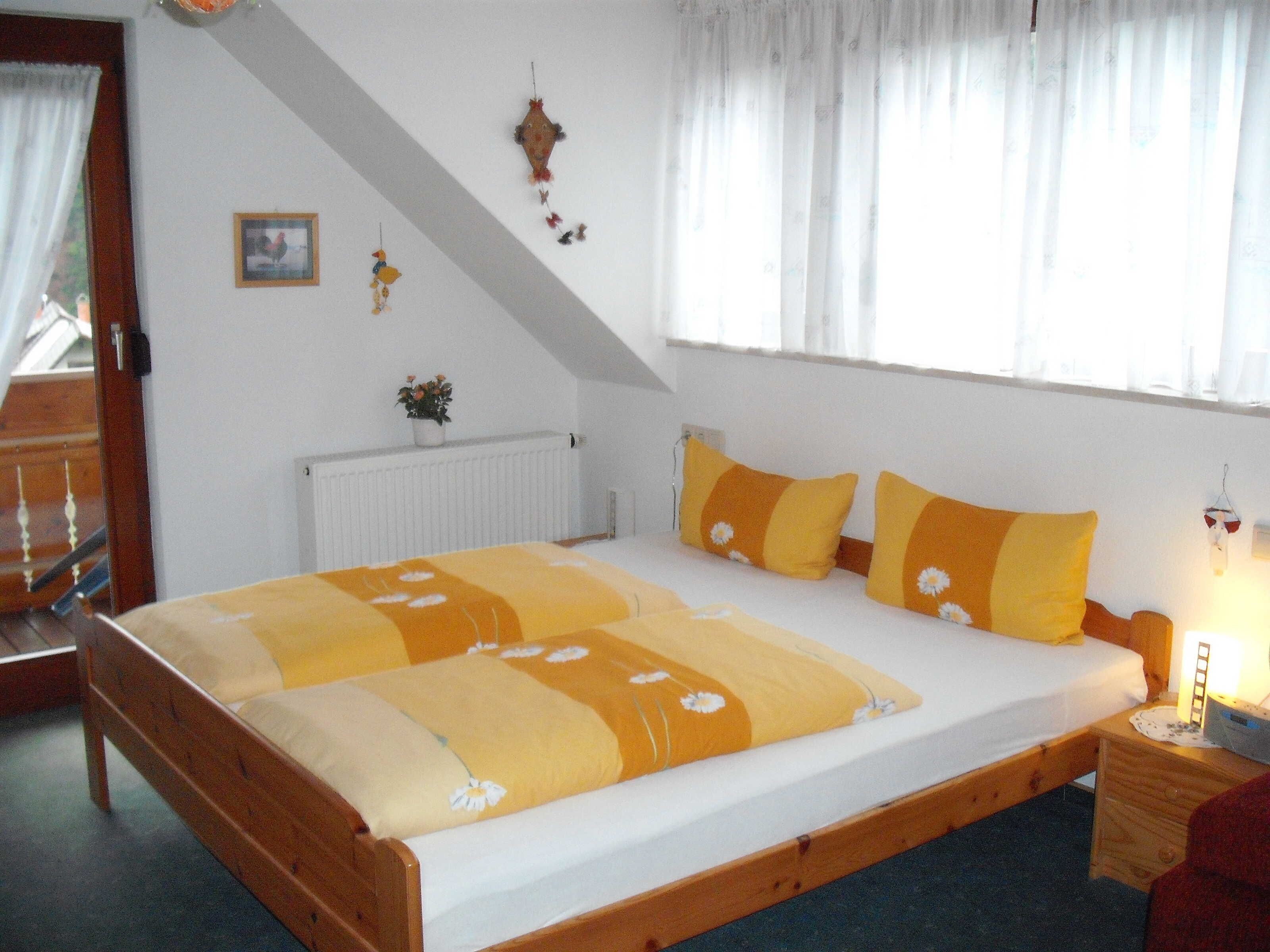 Ein Schlafzimmer mit einem hölzernen Bett, weißen Wänden, einem Fenster mit durchsichtigen Vorhängen, einem Heizkörper und einem kleinen Nachttisch mit Lampe.