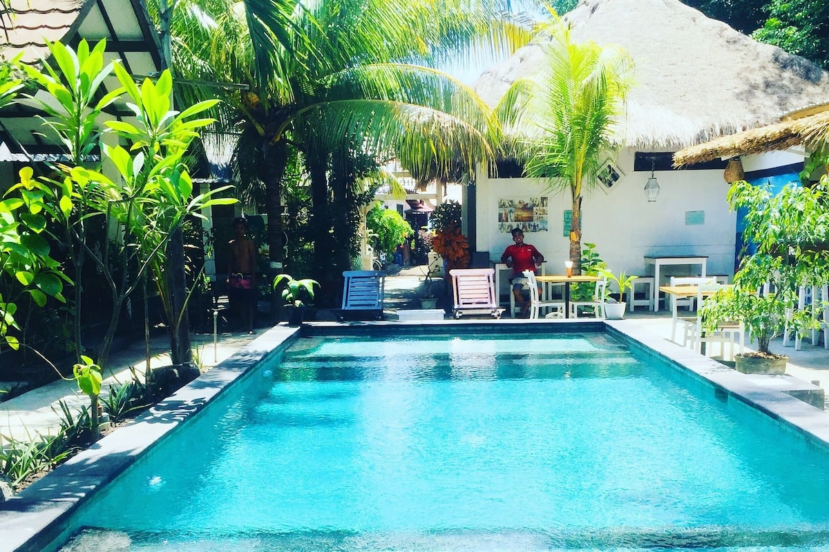 A rectangular swimming pool with a wooden deck in the foreground, surrounded by greenery and a thatched-roof building in the background.