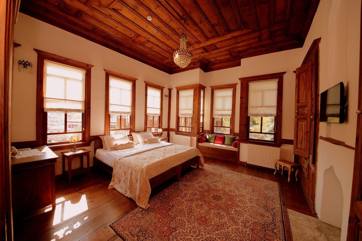 A bedroom with a bed, a wooden ceiling, large windows, a radiator, and a chandelier.