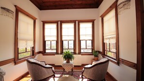 A room with three large windows, wooden flooring, two upholstered chairs, a small table with a potted plant, and a patterned rug. - Karauzumler Konagi (Safranbolu)