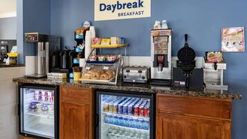 Un área de desayuno con un letrero Daybreak, un refrigerador con bebidas, una cafetera y un fabricante de gofres.