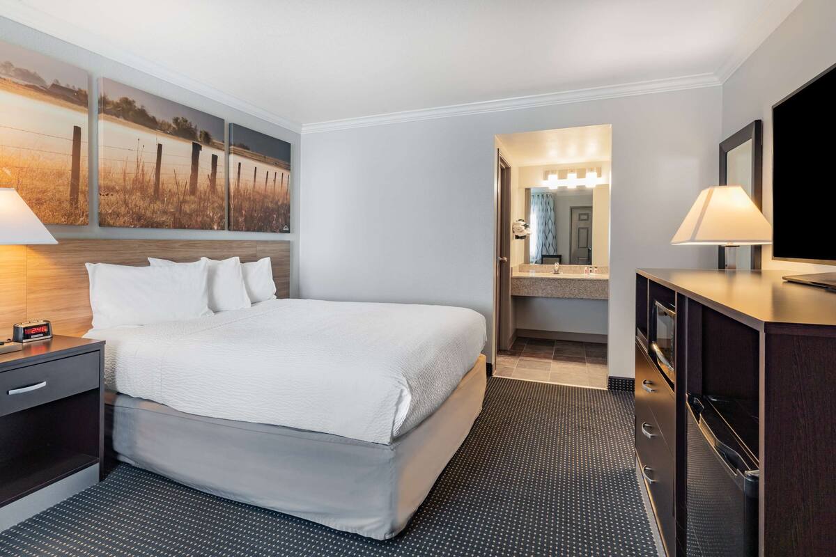 A hotel room with a large bed, a nightstand, a dresser, a TV, and a bathroom visible through an open door.