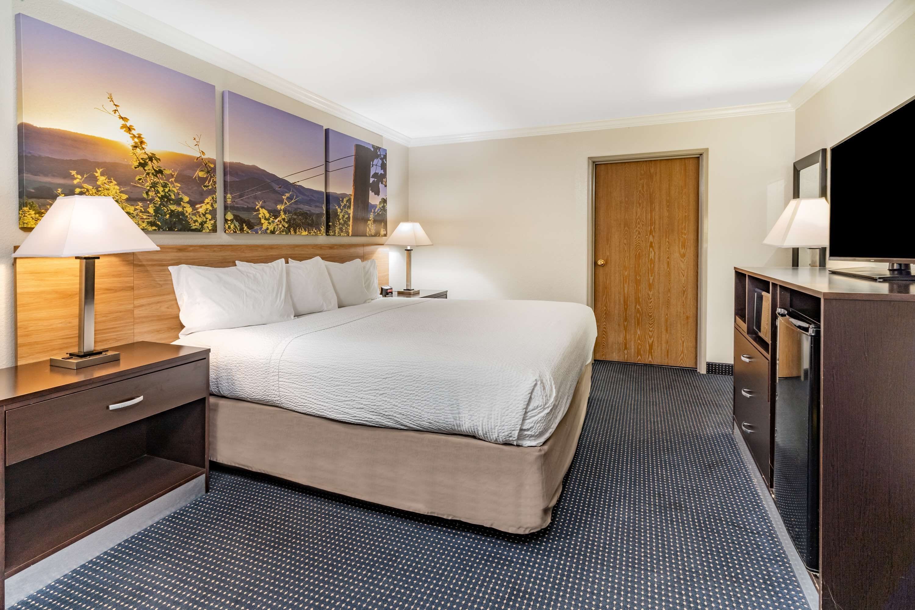 A hotel room with a large bed, two bedside tables with lamps, a flat-screen TV, a wooden wardrobe, and a painting of a sunset on the wall.
