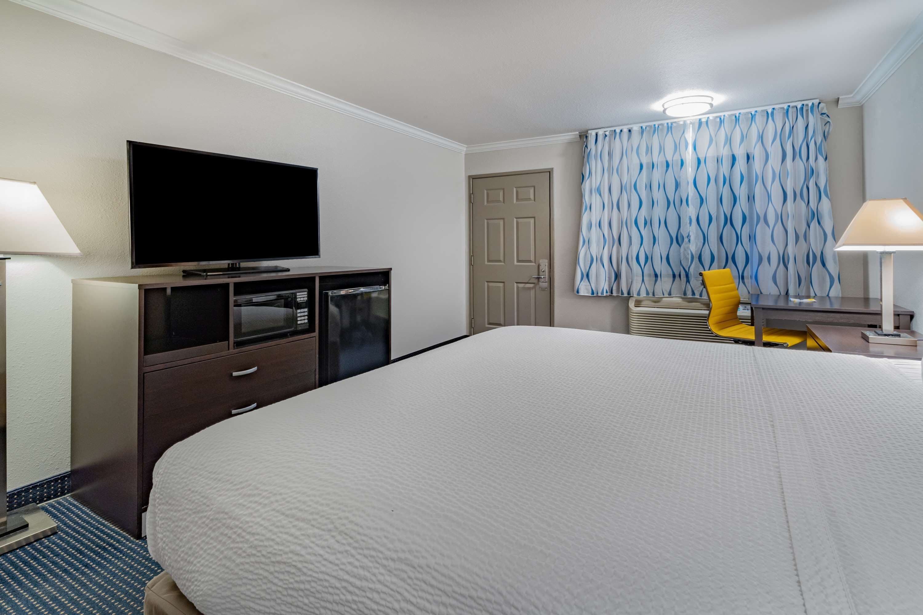 A hotel room with a large bed, a flat-screen TV mounted on the wall, a desk with a chair, and a window with patterned curtains.