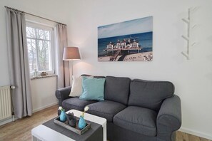 A living room with a grey sofa, a lamp, a picture of a coastal building, and a window with curtains.