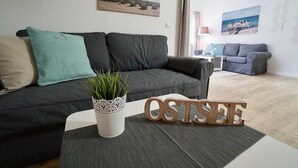 A modern living room with a dark grey sofa, a small white table with a potted plant, and wooden letters spelling "OSTSEE". - 23 Ferienappartement Wittow (with balcony) - Apartment house Haus Arkona (Sellin)
