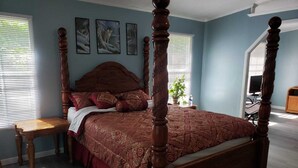 Una cama con dosel con edredón estampado en rojo y dorado, una mesita de noche de madera y una planta en maceta sobre el alféizar.