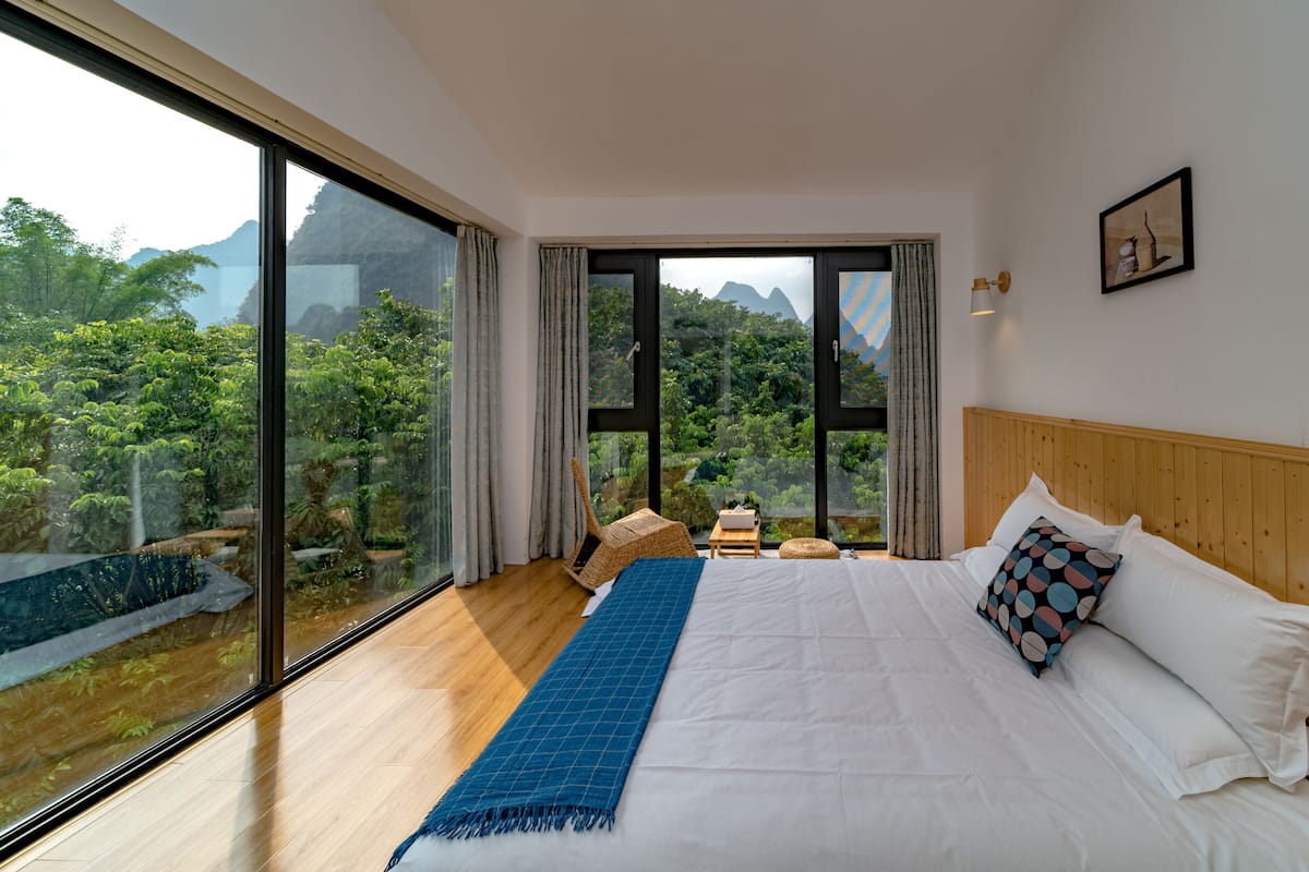 A bedroom with a large window, a bed with white linens, a blue throw blanket, and a wooden headboard.