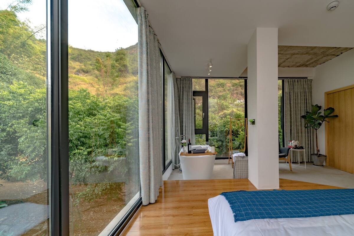 A modern bedroom with a large bed, a bathroom with a bathtub, and a view of a lush green landscape through large windows.