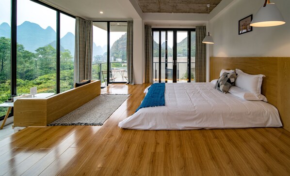 A spacious bedroom with a large bed, a wooden bench, and a view of lush greenery and mountains through large windows.