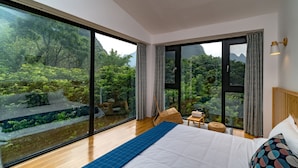 A bedroom with a large bed, a view of a lush garden through floor-to-ceiling windows, and a wooden headboard. - Yangshuo Serene Cove Hotel (Guilin)