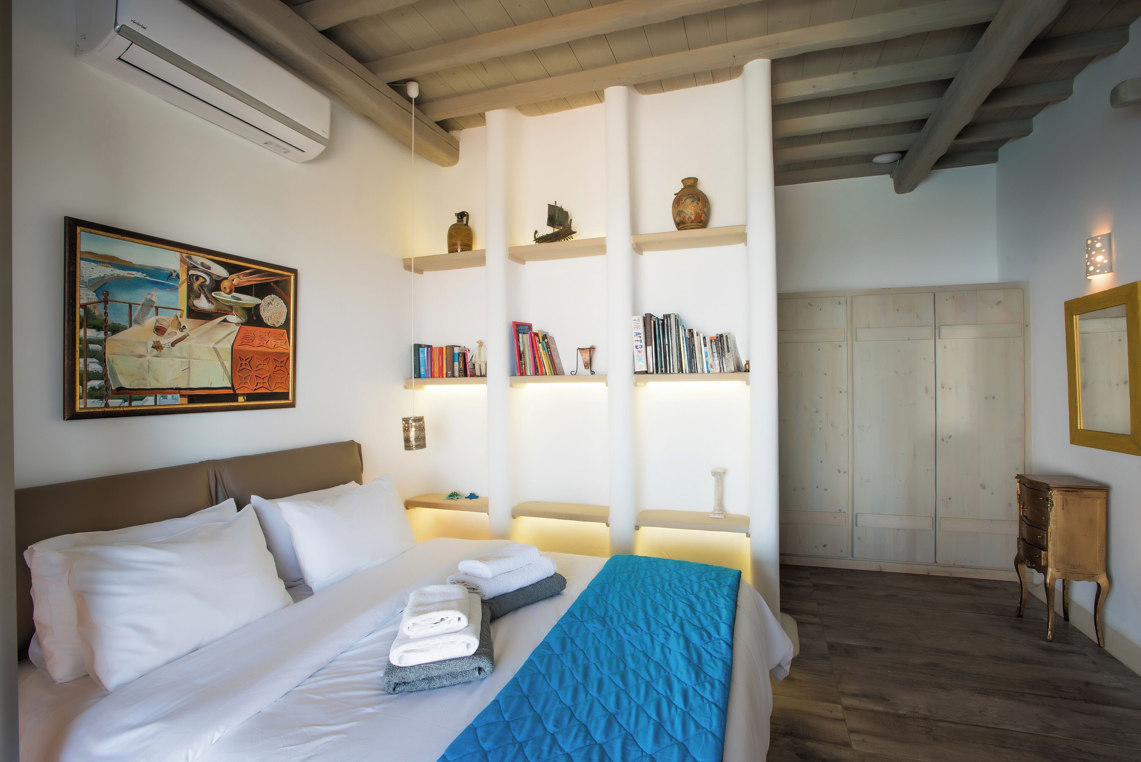 Una habitación de hotel con una cama, un cuadro, estanterías con libros y objetos decorativos, y un armario.