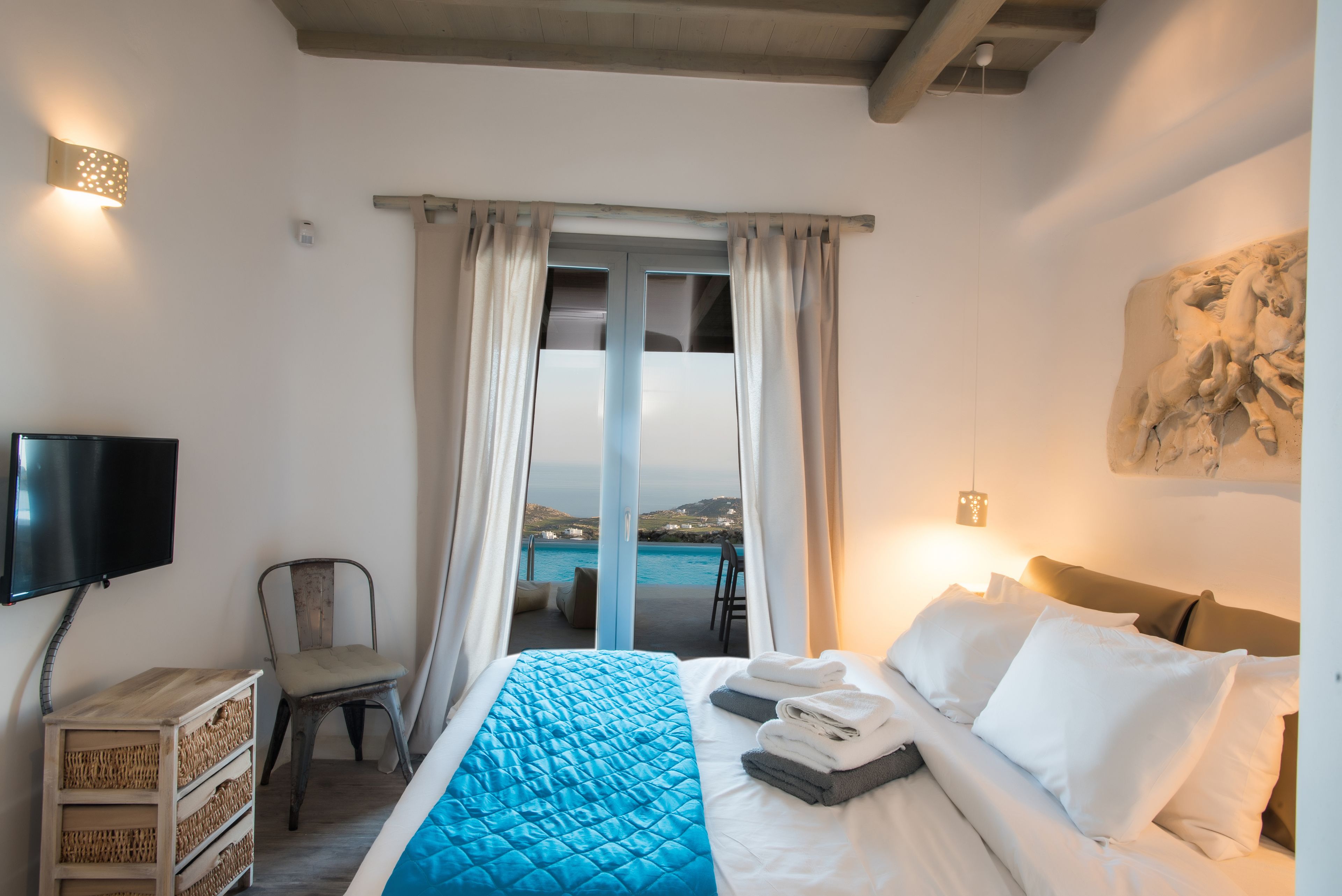 A hotel room with a bed, a chair, a TV, a wall-mounted light, and a view of the sea.