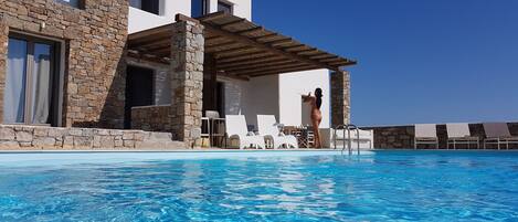 Zona junto a la piscina con una piscina azul cristalina, edificio de piedra y color blanco, y sillas de descanso.