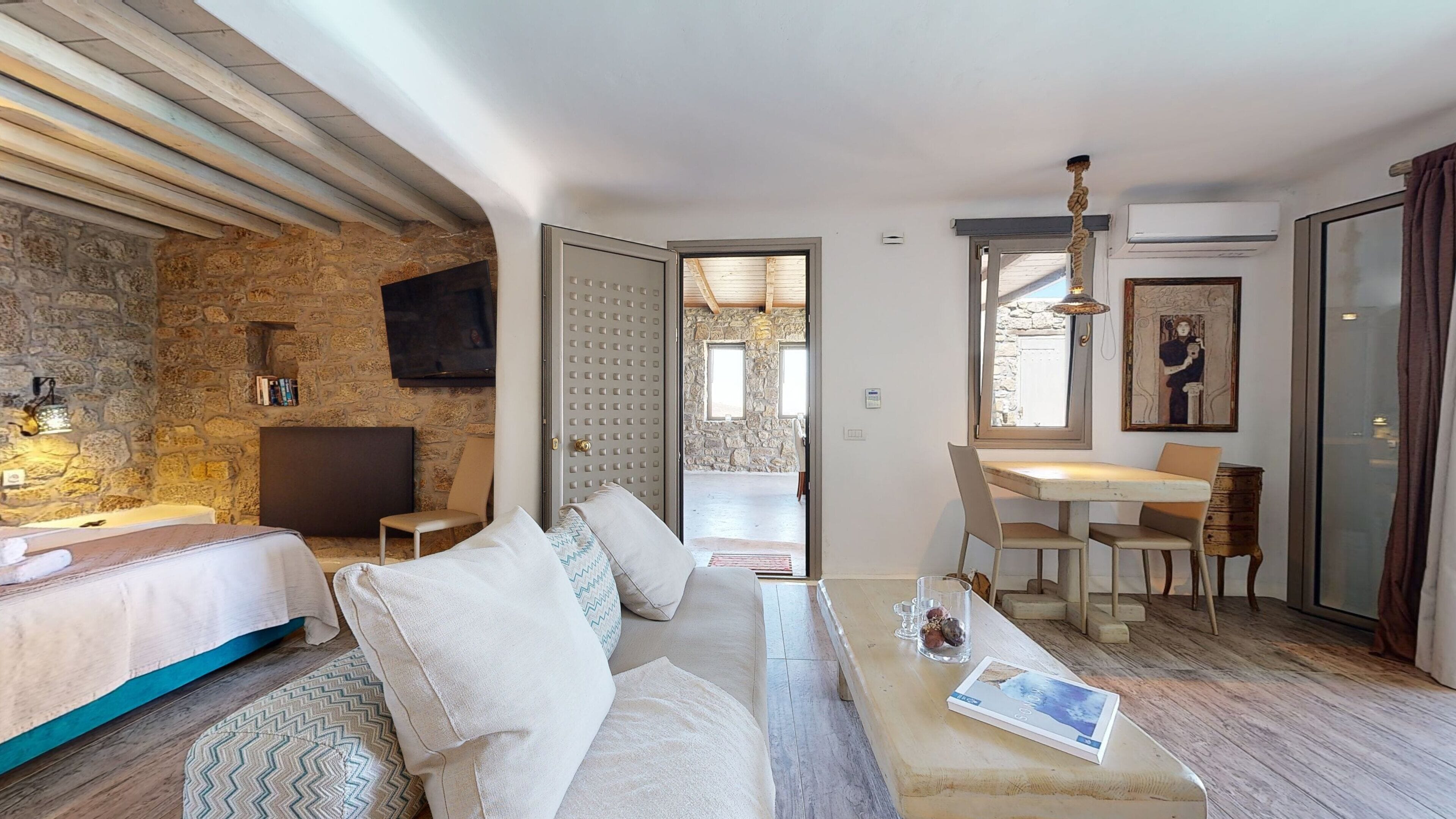 Una camera d'albergo moderna con parete in pietra, un letto, un divano, un'area pranzo con tavolo e sedie, e una TV a parete.