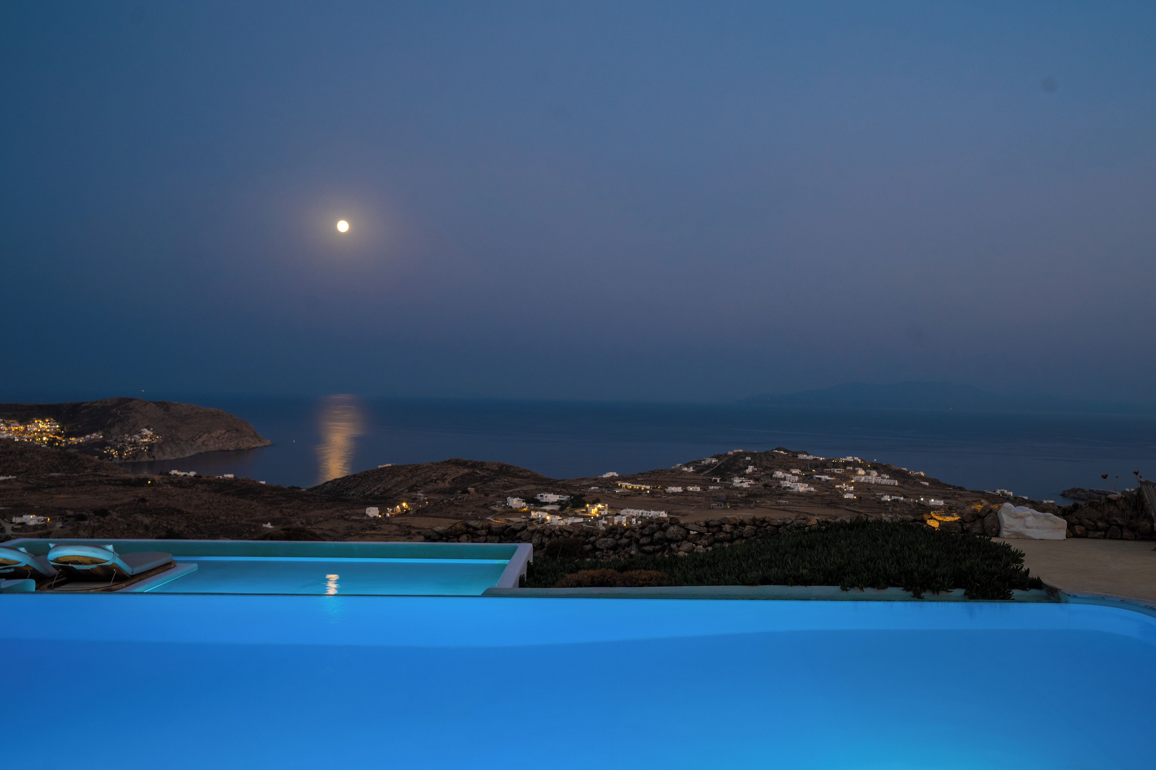 A poolside view with a moonlit night, rocky terrain, and distant buildings.