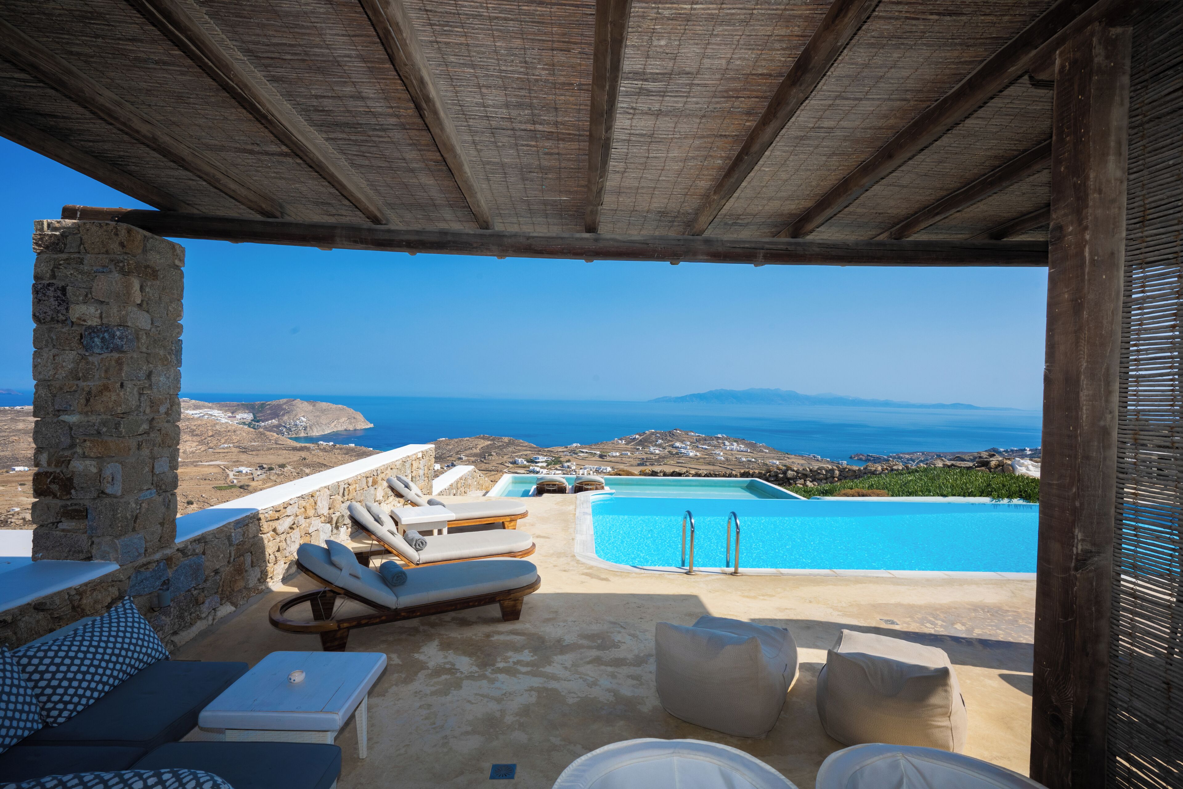 A sunlit outdoor lounge area with a stone pillar, lounge chairs, and a swimming pool.