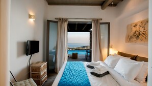 A bedroom with a bed, a TV, a wooden chest, and a view of the sea. - Amallini Suites Mykonos (Mykonos)