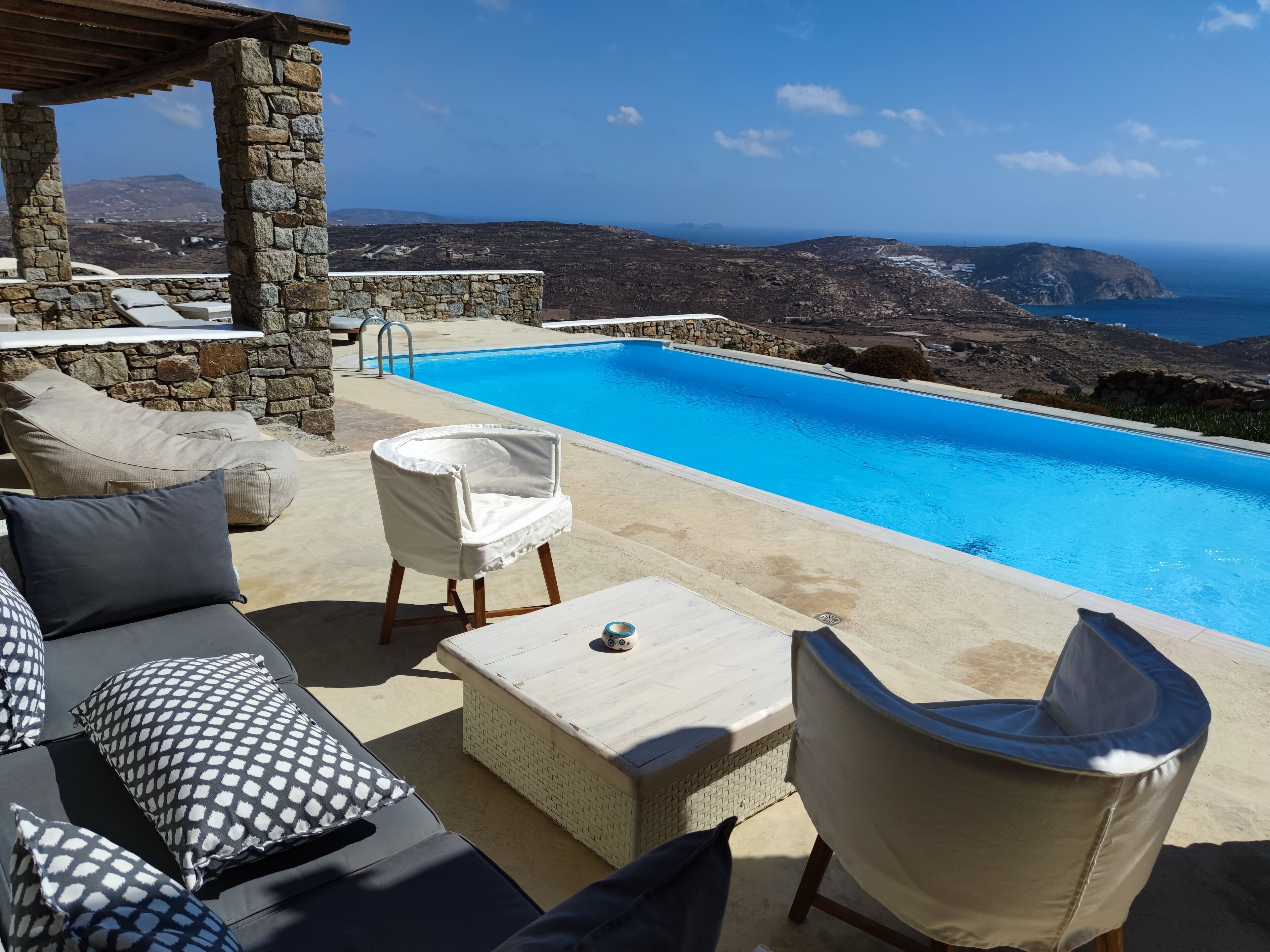 Un espace aménagé au bord de la piscine, avec des piliers en pierre, une chaise blanche, un canapé agrémenté de coussins à motifs et une vue sur la mer et les montagnes.