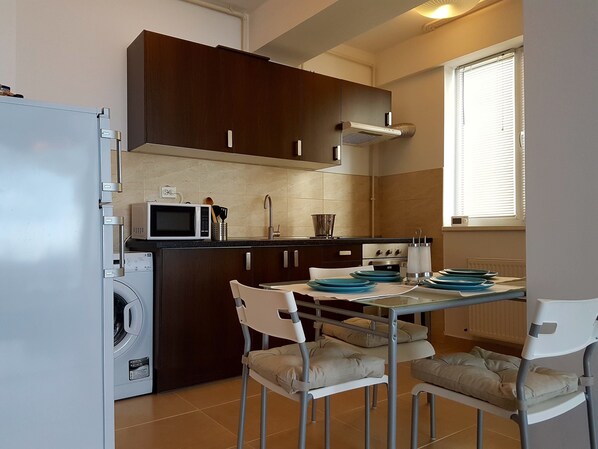A compact kitchen with a built-in washing machine, a microwave, and a stove. There are white chairs around a small table with blue plates. - Apartment Summerland Mamaia (Constanta)