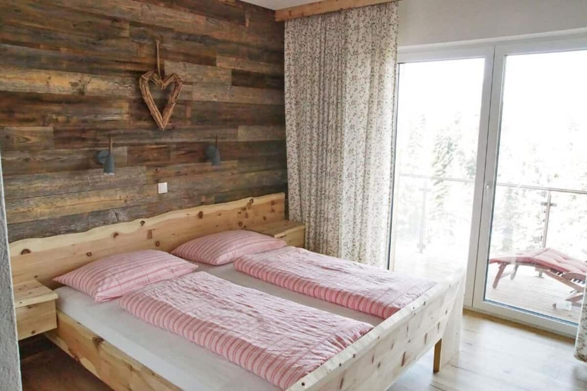 Ein Schlafzimmer mit hölzernem Kopfteil, zwei Betten mit rosa gestreifter Bettwäsche, einer herzförmigen Dekoration und Blick auf einen Balkon mit Gartenmöbeln.