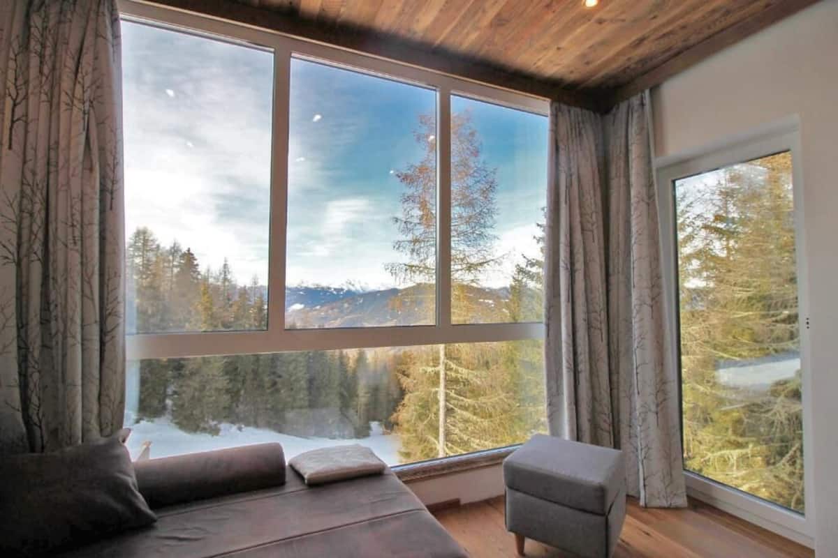 Ein Zimmer mit einem großen Fenster, das einen Blick auf eine verschneite Landschaft und Bäume bietet. Im Zimmer befinden sich eine Couch und ein Sessel.