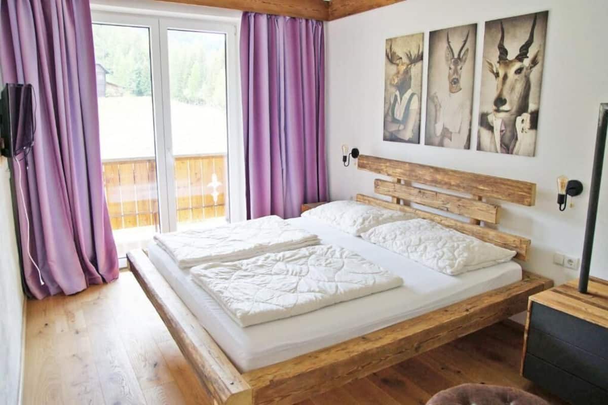 Ein hölzernes Bett mit weißer Bettwäsche in einem Raum mit violetten Vorhängen und einer Holzbalkendecke.