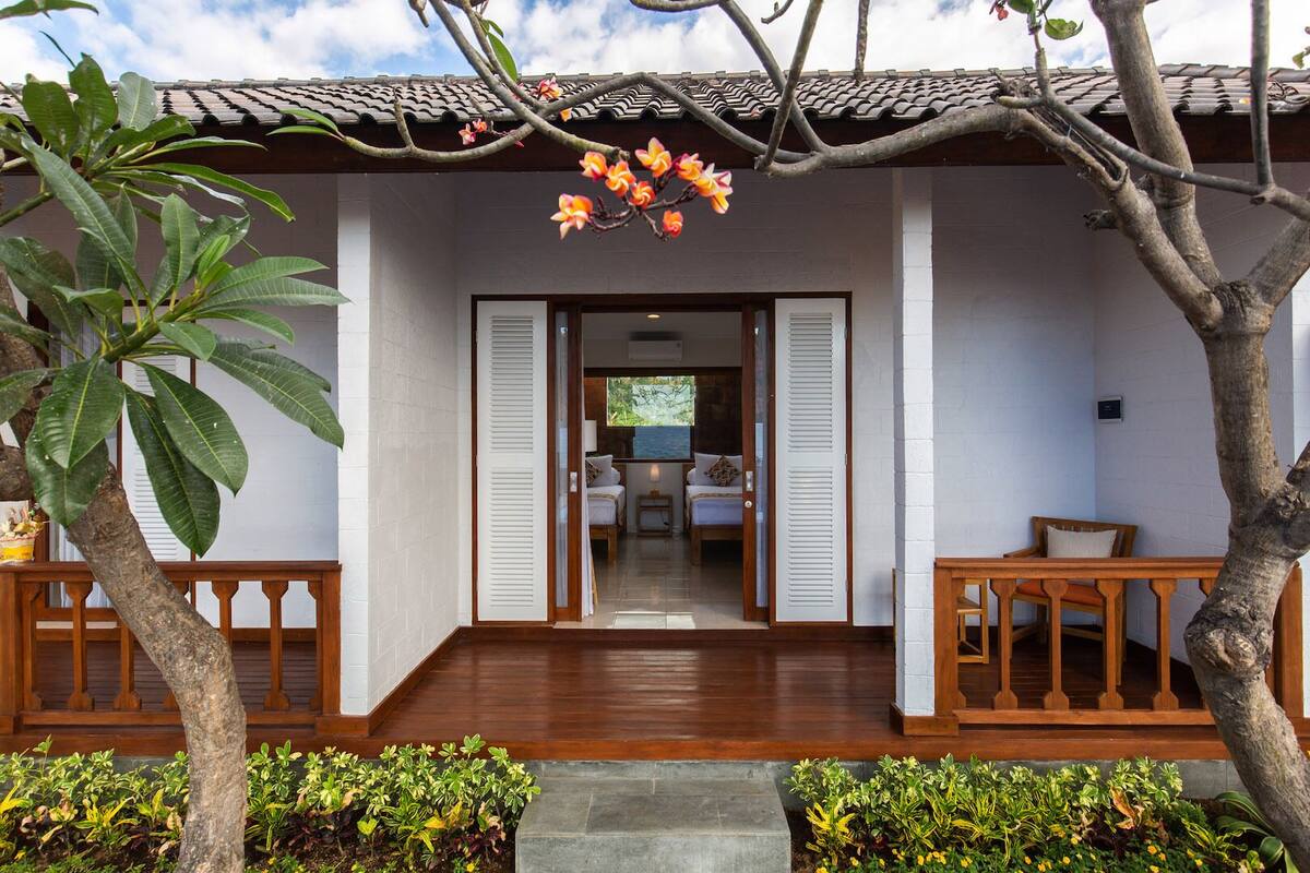 A traditional-style building with a wooden deck, a small garden area with plants, and a view into a room with a bed and a desk.
