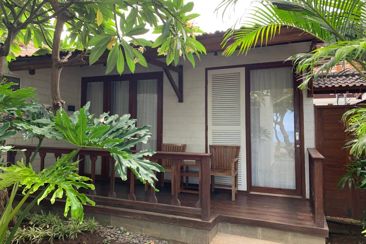 A wooden deck with a bench, surrounded by lush greenery and a building with large windows and a door.