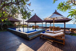 Terraza de madera con piscina, sillas de descanso y sombrillas con vistas al mar.