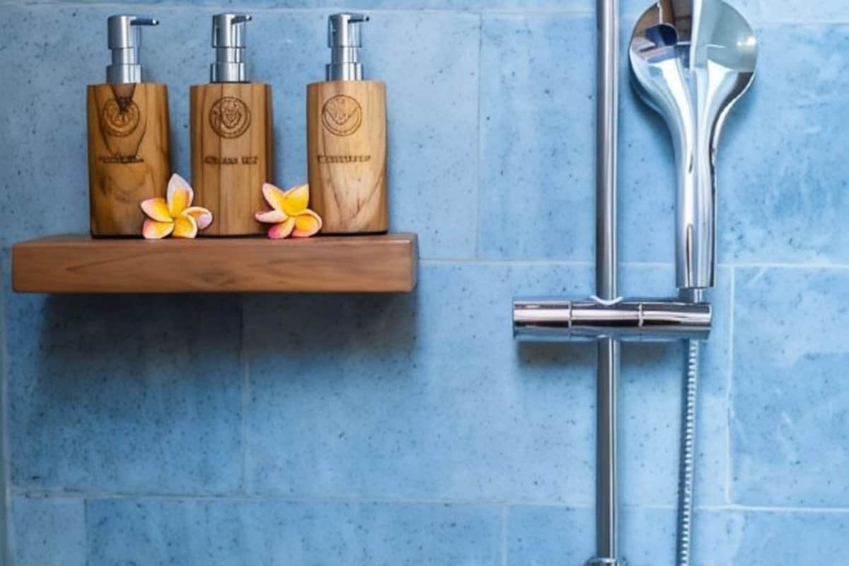 A bathroom with a wooden shelf holding three bottles, a showerhead, and a faucet.