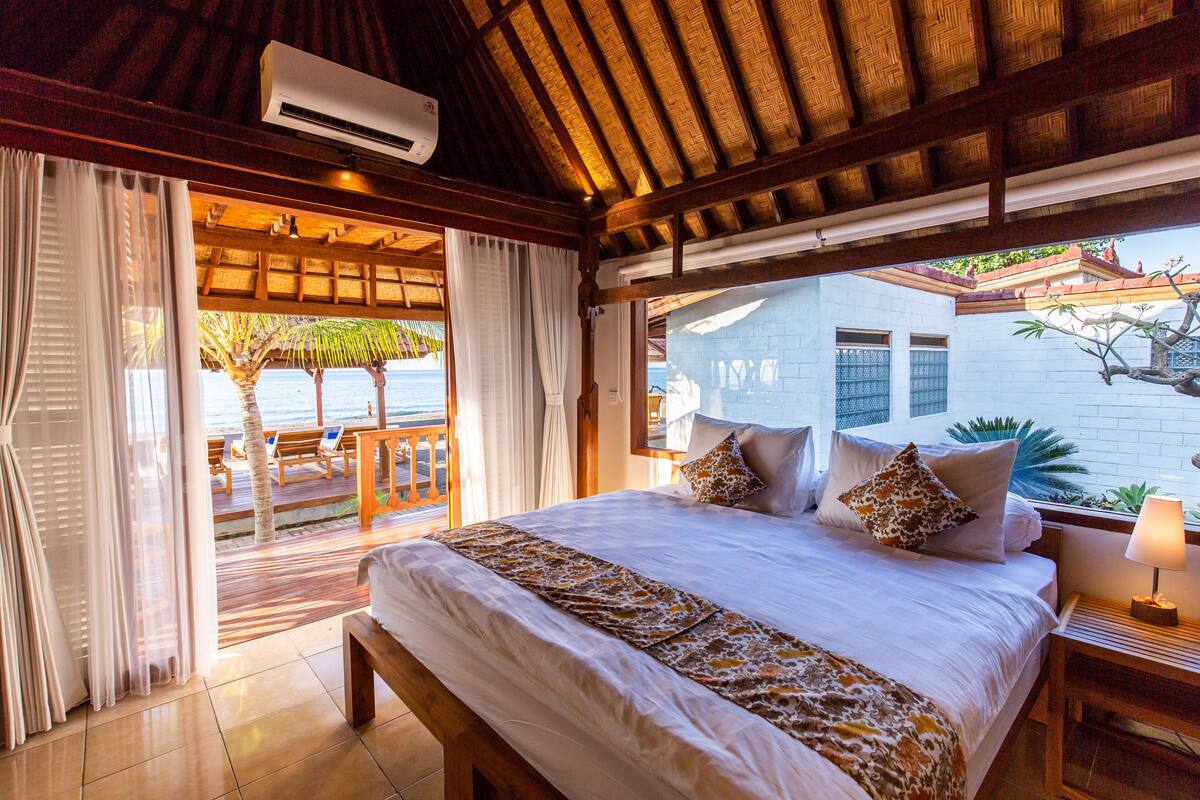 A bedroom with a large bed, a wooden headboard, a nightstand with a lamp, and a view of the ocean and palm trees through a sliding glass door.