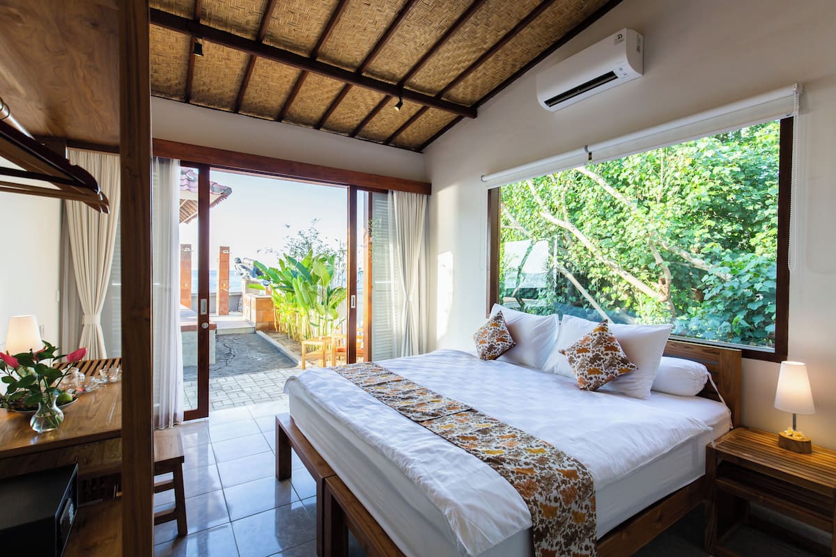 A bedroom with a large bed, a wooden headboard, a nightstand with a lamp, and a view of greenery outside.