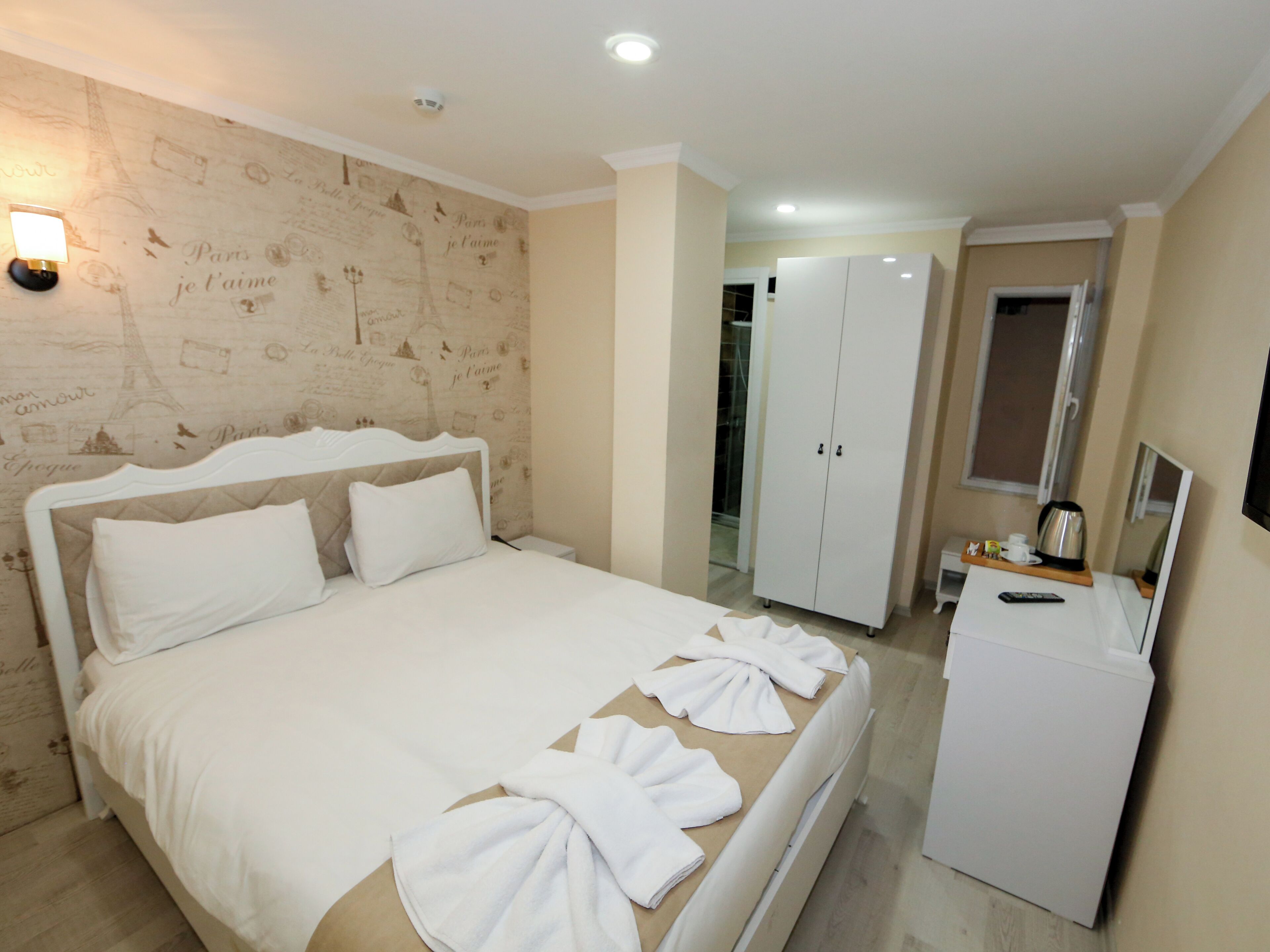 A hotel room with a bed, a wardrobe, a desk with a kettle, and a wall with Paris-themed wallpaper.