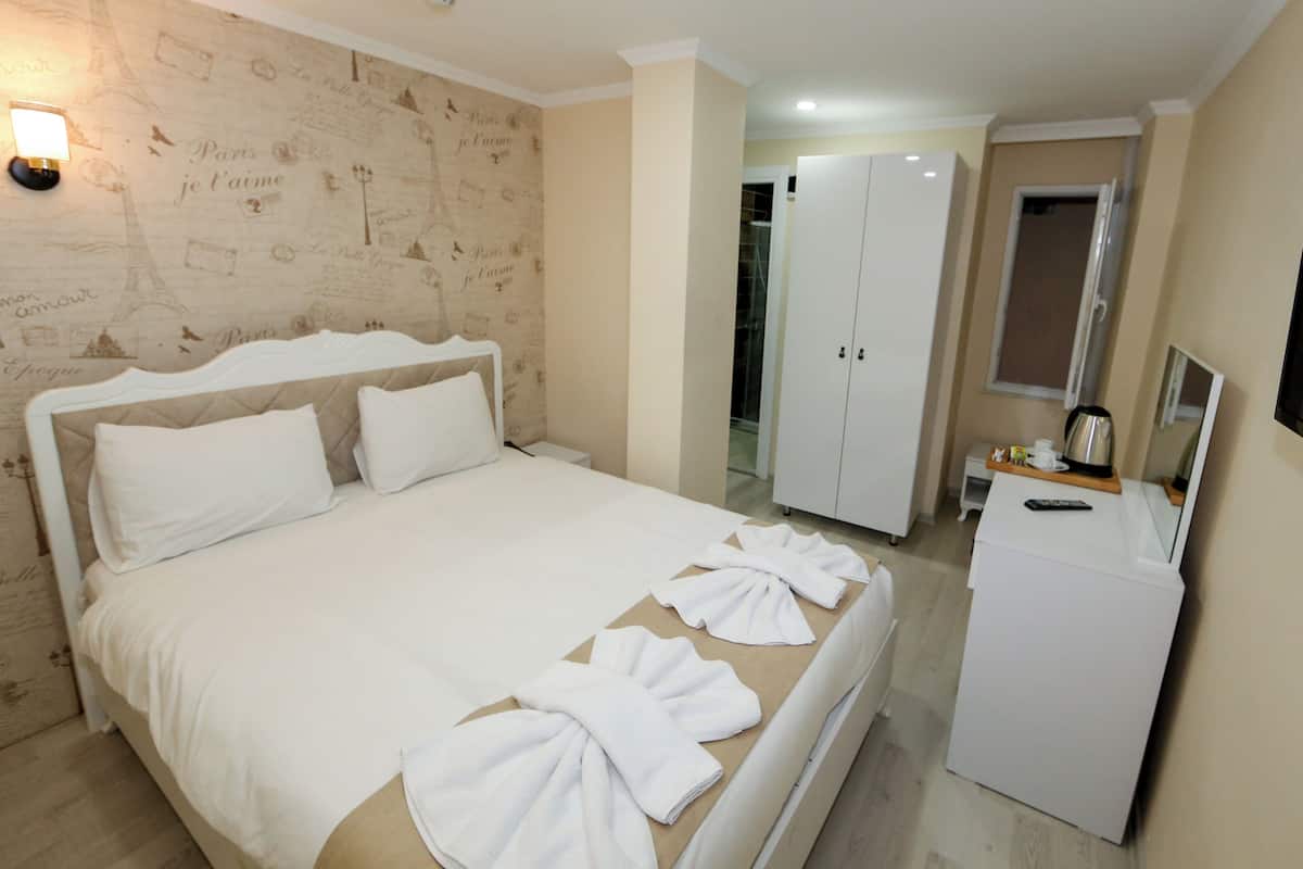 A hotel room with a bed, a wardrobe, a desk with a kettle, and a wall with Paris-themed wallpaper.