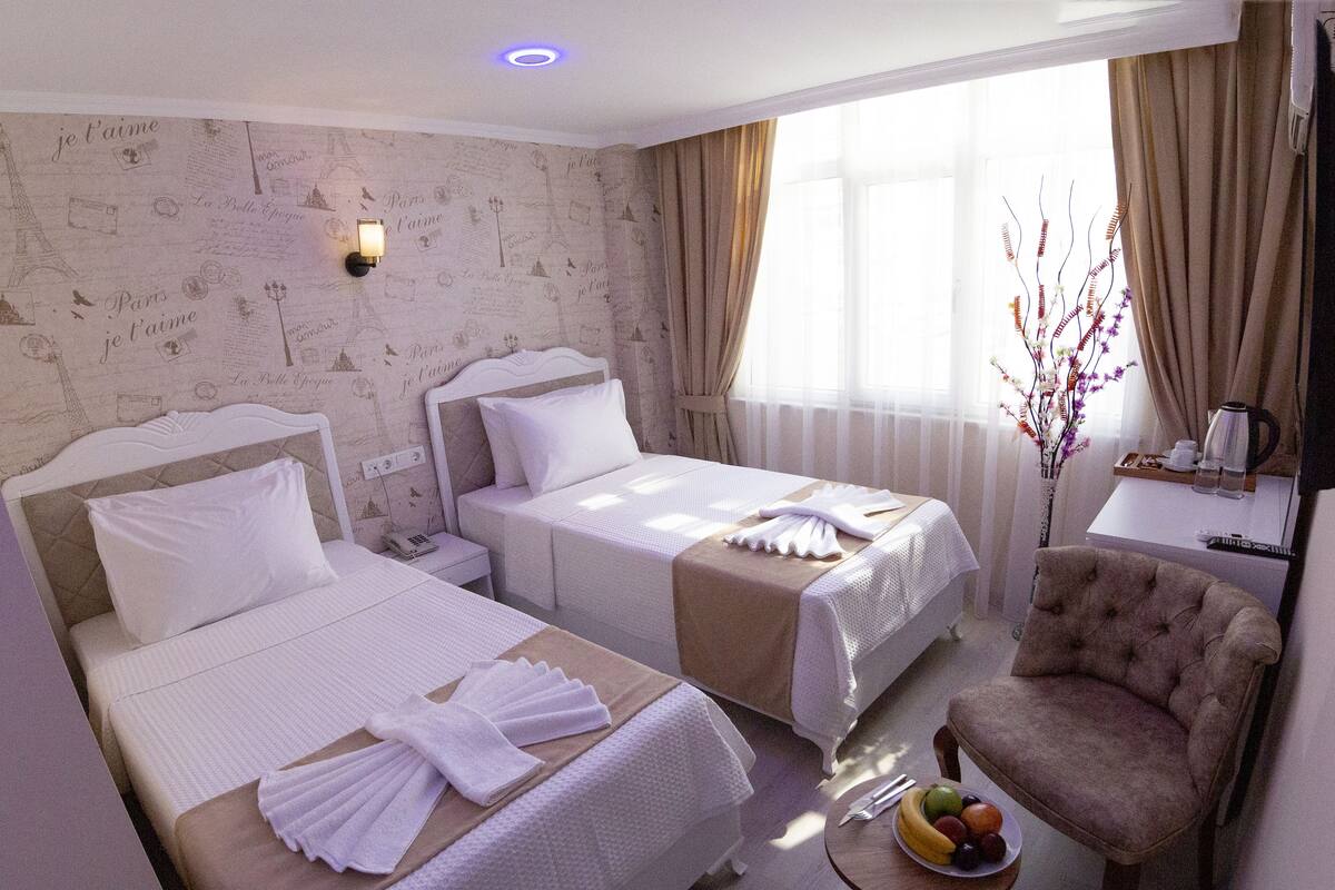 A hotel room with two beds, a chair, a small table with fruit, and a window with curtains.