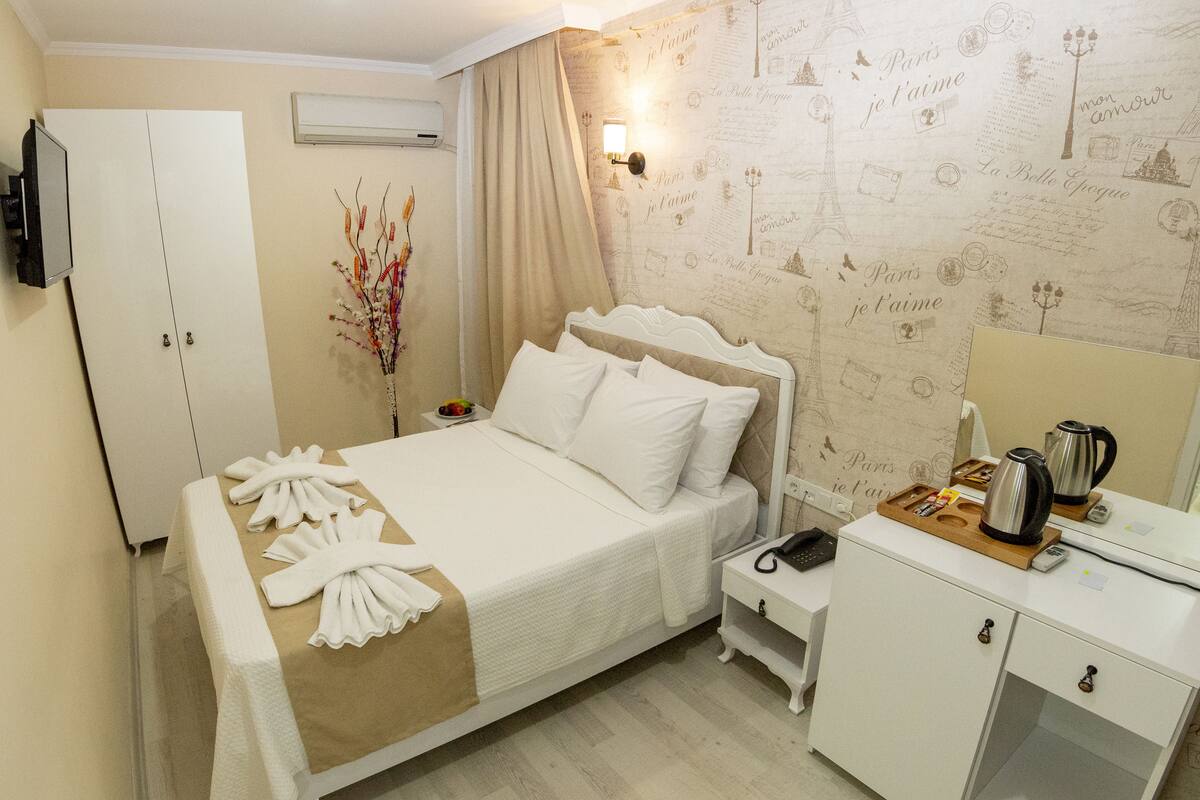 A hotel room with a bed, a wardrobe, a bedside table with a lamp, a mirror, and a wall with Paris-themed wallpaper.