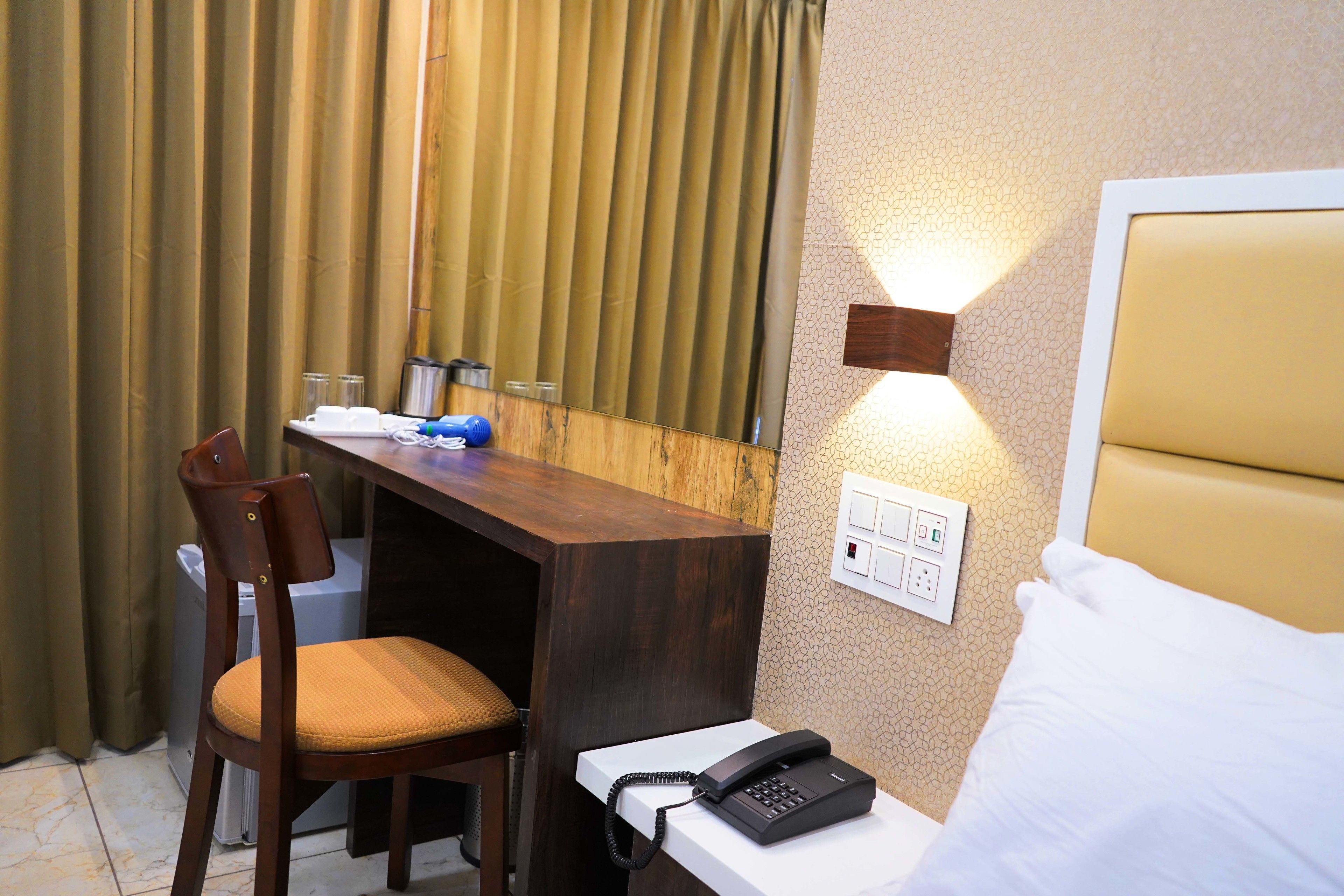 A hotel room with a wooden desk, a chair, a bed with white bedding, a bedside phone, and a wall-mounted light.