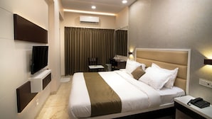 A modern hotel room with a large bed, a flat-screen TV mounted on the wall, a desk with a chair, and an air conditioning unit. - Hotel Royal Treat (Surat)