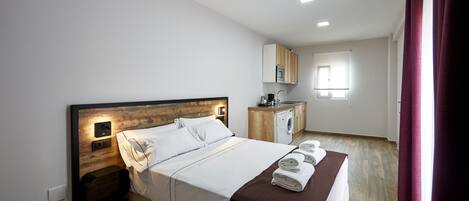 A modern hotel room with a wooden headboard, a bed with white linens, a bedside table, a built-in microwave, a small kitchen area, and a window with curtains.