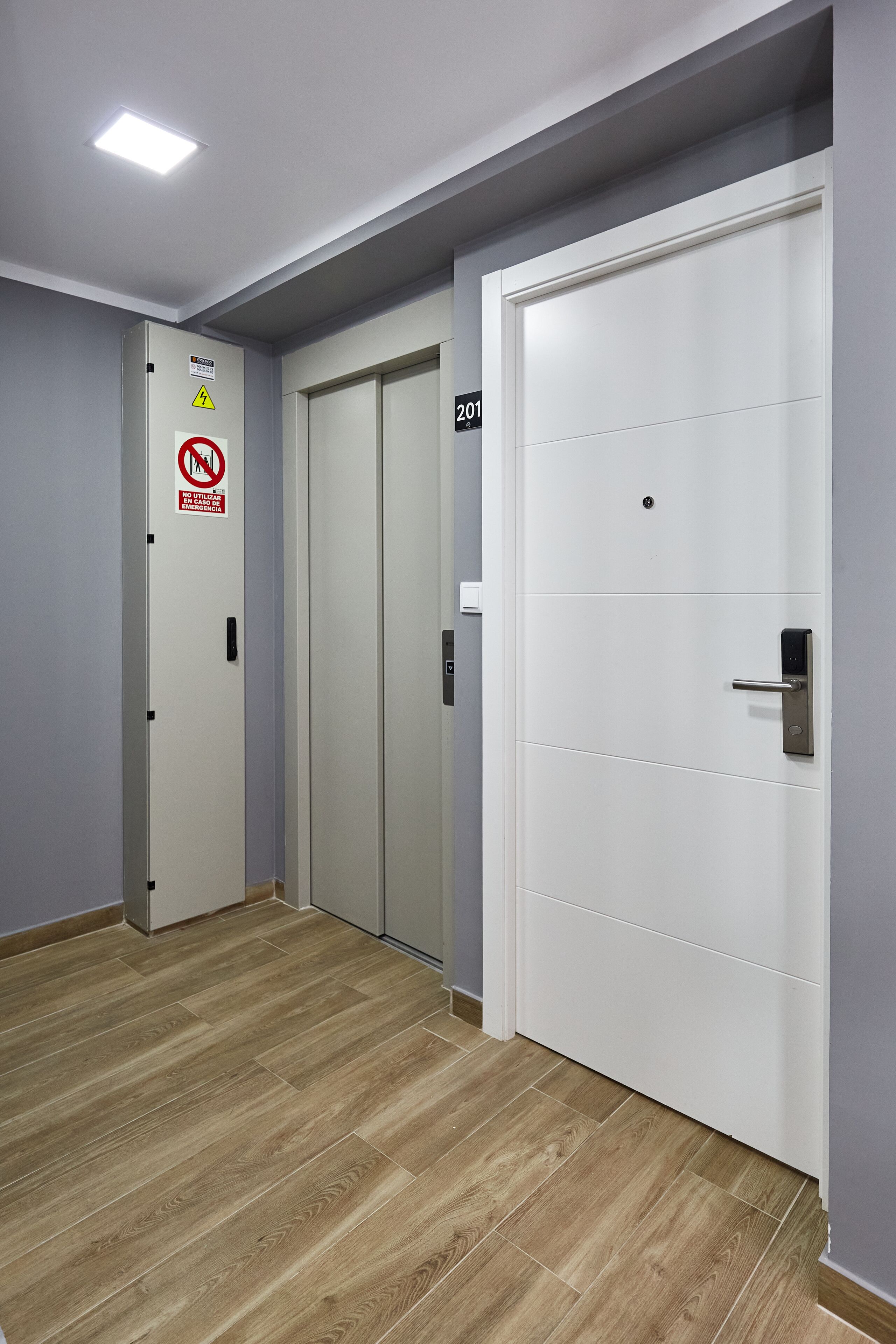 A hallway with a wooden floor, a white door with a number plate, and a no smoking sign.
