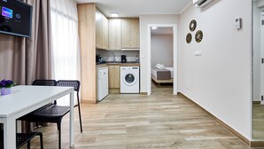 A compact living space with a kitchenette, a washing machine, a dining area with a table and chairs, and a small bedroom with a bed and a bedside table. - GBH Hotel-Apartamentos Caballito de Mar (Benidorm)