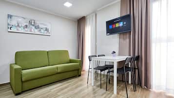 Un salon moderne avec un canapé vert, une table à manger blanche et quatre chaises, un téléviseur à écran plat fixé au mur et un tableau représentant un paysage urbain accroché au mur.